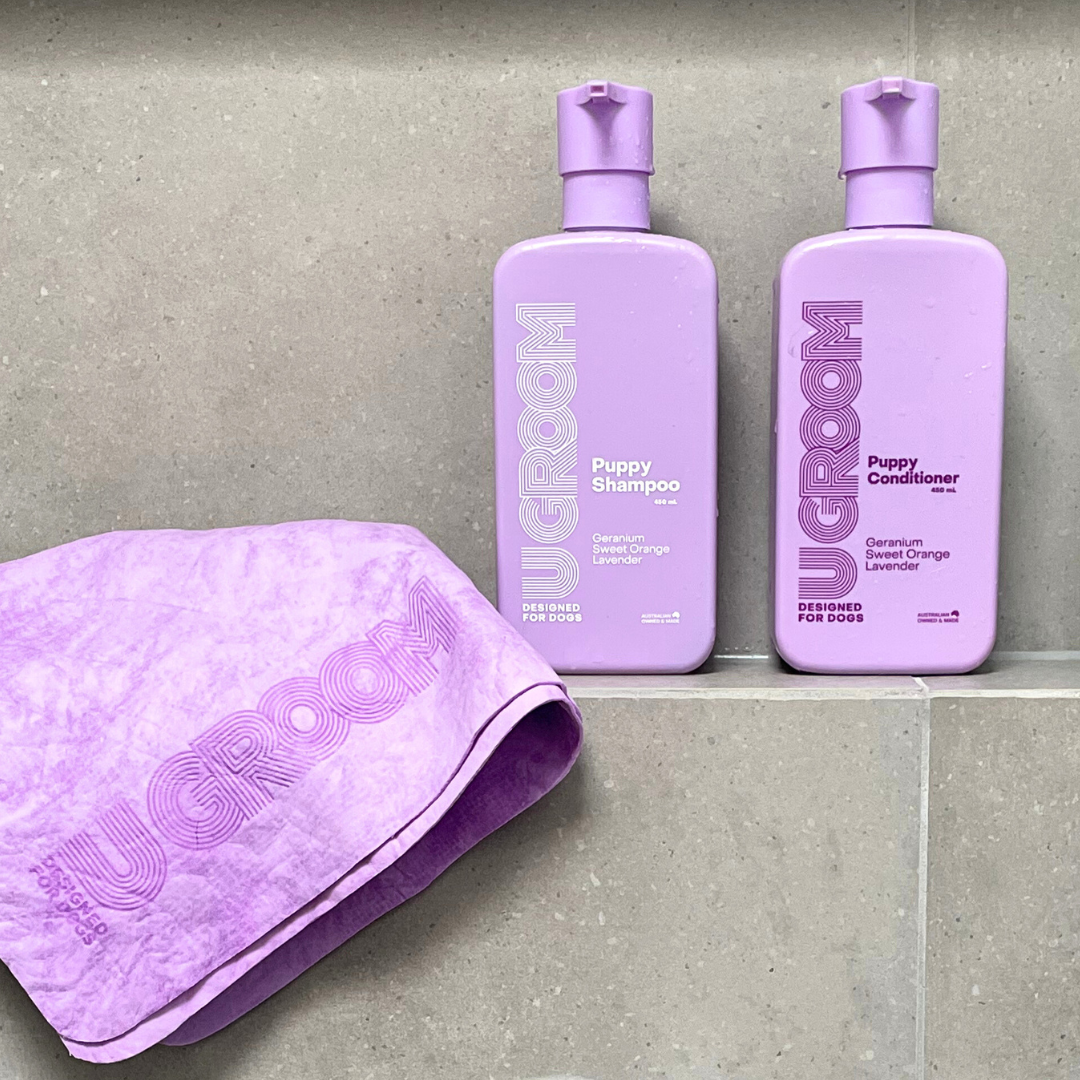 Two purple bottles of UGROOM puppy shampoo and conditioner with a matching towel on a gray surface.